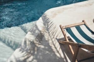 A striped deckchair sits on a sunlit poolside patio, where you can simply be, palm tree shadows dancing across the concrete next to the clear blue water.