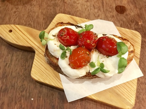 A slice of toasted bread topped with fresh mozzarella, roasted cherry tomatoes, and green herbs, served on a small wooden board with a piece of greaseproof paper—perfect for the Super Lunch Menu at Jamie’s Italian.