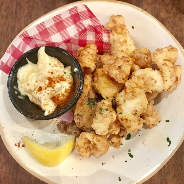 A plate of golden fried calamari from Jamie's Italian, served with a lemon wedge and a black cup of creamy dipping sauce sprinkled with paprika, all on a white plate lined with red chequered paper—a perfect choice from the Super Lunch Menu.