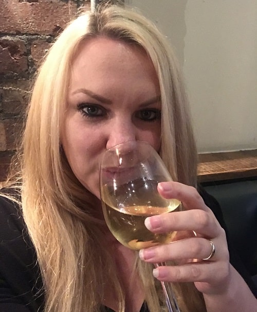 A woman with long blonde hair is sitting indoors at Jamie’s Italian, holding and sipping from a glass of white wine. She is looking at the camera and wearing a black top, with a visible brick wall in the background—perfect for a review of the Super Lunch Menu.