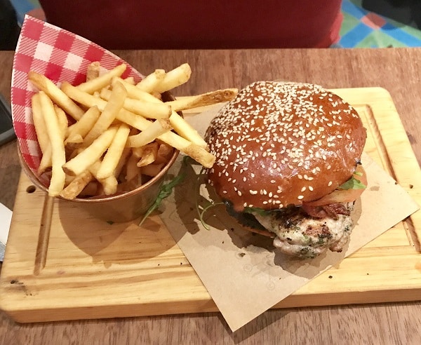 A sesame seed burger bun filled with lettuce, bacon, and cheese, served on a wooden board with golden chips in a metal container lined with red and white chequered paper—a tasty pick from the Super Lunch Menu at Jamie’s Italian.
