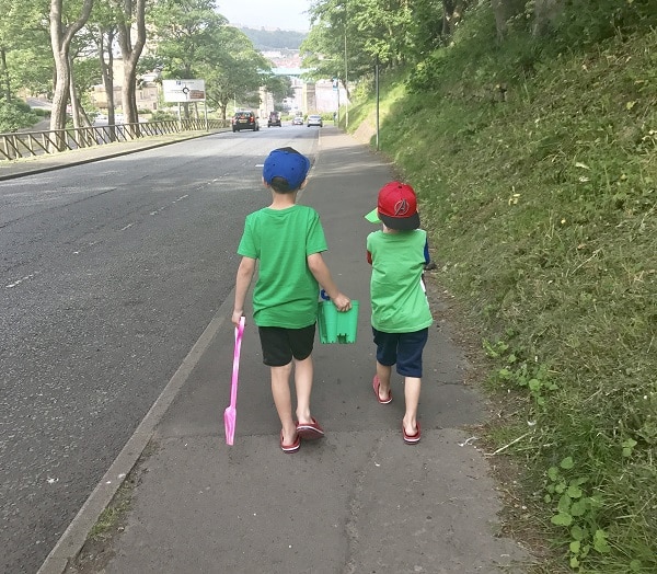 Two children wearing green shirts and red caps walk on a pavement beside a road, showing alternative ways to get kids active. One holds a pink spade, the other carries a green object. Trees and grass line the path, with buildings in the distance.