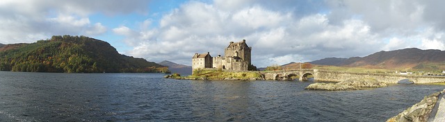 A stone castle stands on a small island connected to the shore by an arched stone bridge, surrounded by water and rolling hills under a partly cloudy sky—a perfect scene for travel in the UK.