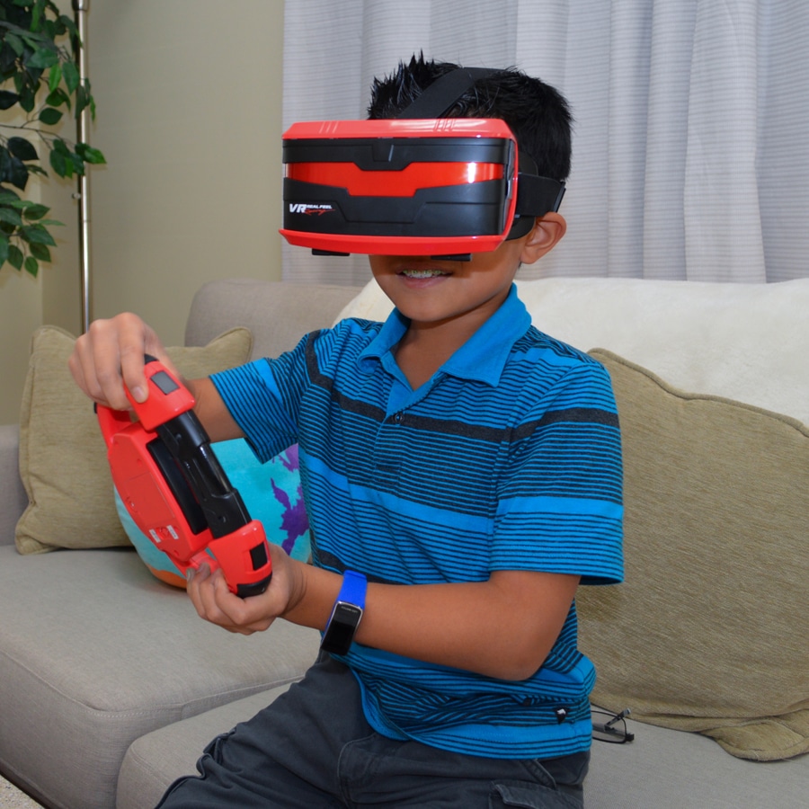 A smiling boy wearing a red and black VR headset from VR Real Feel Racing sits on a beige sofa, holding a red games controller. He is dressed in a blue striped polo shirt and grey shorts, ready to win in the world of virtual reality systems.
