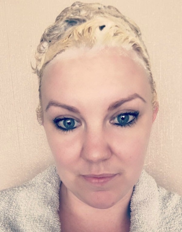 A woman with fair skin and blue eyes looks at the camera, her hair covered in bleach or hair dye—showcasing hair essentials for a salon look at home, while she wears a grey towel around her shoulders against a neutral background.