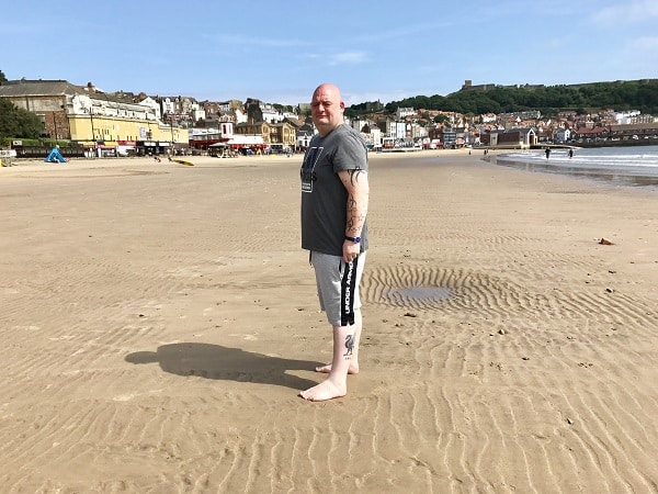 A man stands barefoot on a sandy beach in a grey t-shirt and shorts, looking Summer Ready. Buildings and greenery are visible in the background under a blue sky.