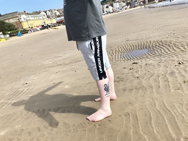 Person standing barefoot on wet sand at the beach, wearing grey Under Armour shorts and looking summer-ready. Their lower leg sports a bird tattoo, with buildings and more beach stretching out in the background.