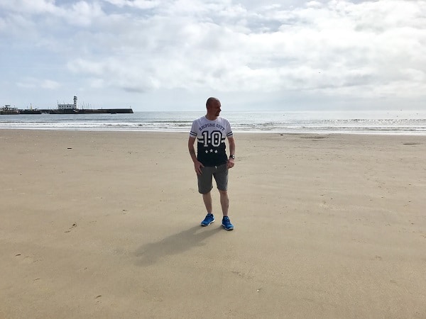 A man in a white T-shirt, grey shorts, and blue trainers stands alone on a wide, sandy beach with the sea and a pier behind him under a partly cloudy sky—summer ready in stylish Jacamo attire.