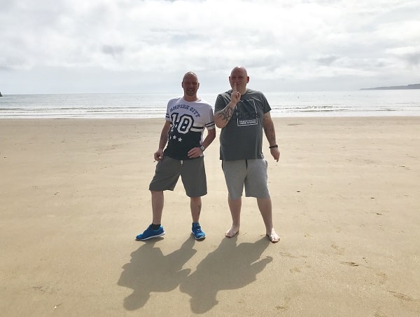 Two men stand on a sandy beach with the sea and cloudy sky in the background, showcasing Jacamo summer fashion. One poses with hands on hips whilst the other gestures with a finger over his lips. Both wear stylish shorts and casual T-shirts.