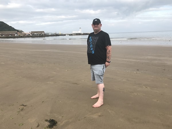 A man in a black cap, black Adidas t-shirt, and grey shorts stands barefoot on a sandy beach beneath a cloudy sky, with city buildings and the sea behind him—looking summer ready in his Jacamo style.
