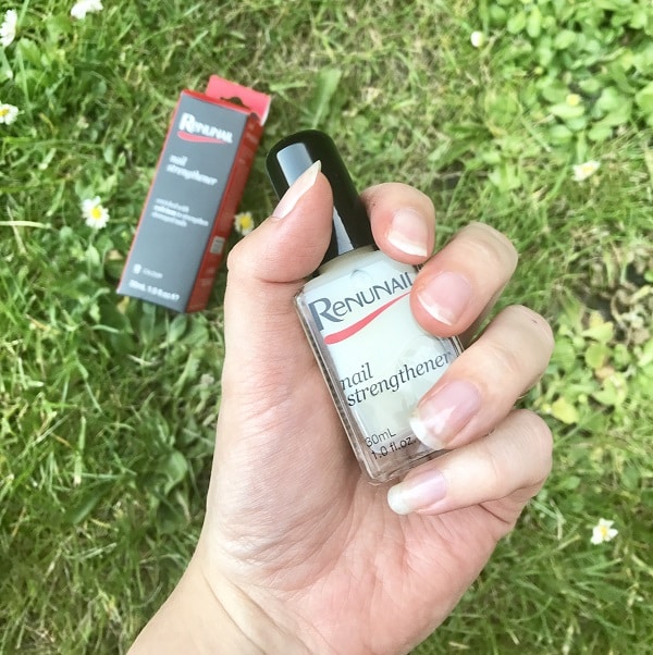 A hand holding a bottle of Renunail nail strengthener with neatly manicured nails, achieving a salon look at home; the product box and grass with small flowers are visible in the background.