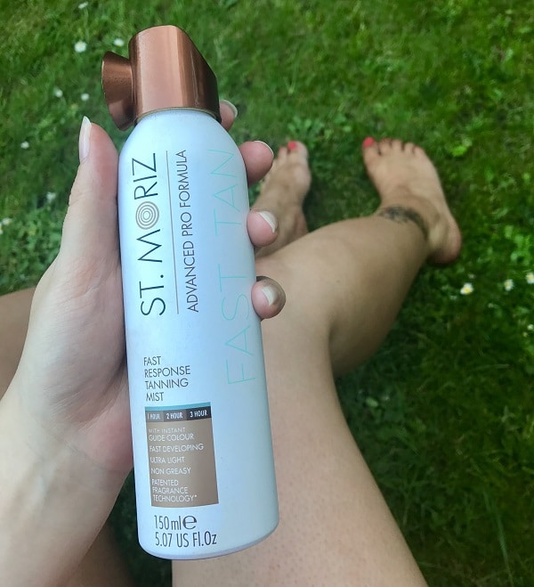 A person holds a can of St. Moriz Advanced Pro Formula Fast Tan Tanning Mist—one of the ultimate body essentials—for a flawless glow. Their legs and grass are visible in the background; the can is white with a bronze cap and blue text.