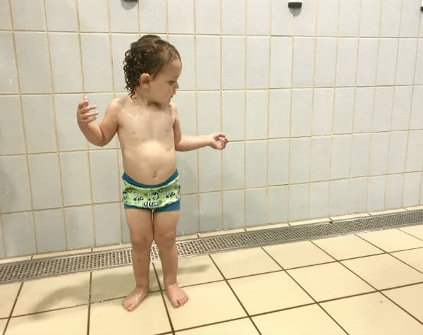 A young child with wet hair stands on tiled flooring by a tiled wall, wearing only a Splash About swim nappy. The child looks to the side with arms slightly raised, suggesting a swimming pool or shower area.