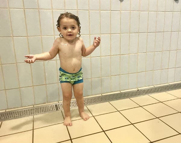 A young child in swim trunks and a Gecko Happy Nappy reusable swim nappy stands barefoot on a tiled floor, wet hair and soap bubbles on their shoulders, looking at the camera with arms slightly raised—perfect for Splash About moments.