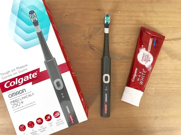 A Colgate ProClinical 250+ electric toothbrush is shown with its box on a wooden surface, next to a tube of Colgate Optic White toothpaste—perfect for adding to your collection of body essentials for that salon look at home.