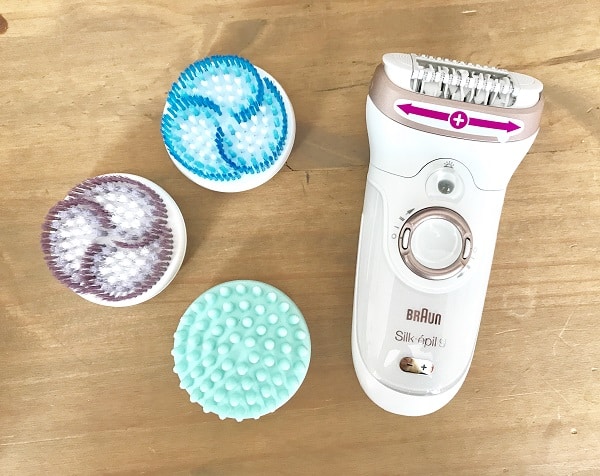 A white Braun Silk-épil 9 epilator with three interchangeable brush heads—blue, purple, and green—placed on a wooden surface. Pink arrows on the epilator show the direction of movement for your hair essentials routine.