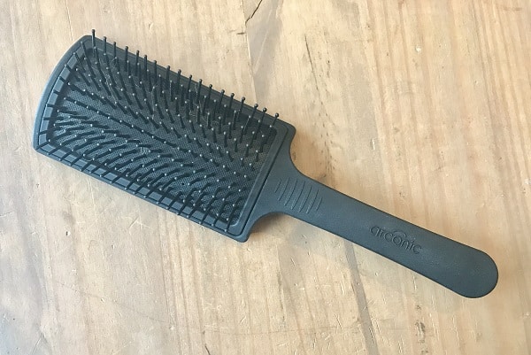 A black rectangular paddle hairbrush with plastic bristles lies on a wooden surface. The word "arconic" is embossed on the handle—an essential tool for achieving a salon look at home.