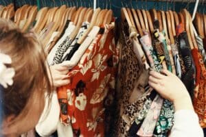 A person browses through a rail of colourful, patterned clothing on wooden hangers in a shop, searching for pieces to create a classic look. Only their hands and part of their head are visible as they explore the fashion options.