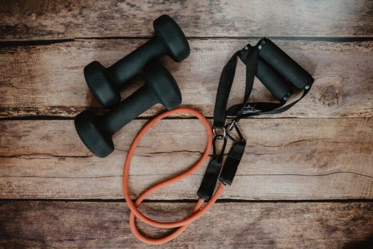 Two black dumbbells and an orange resistance band with black handles sit on a wooden surface, perfect for carving out some me time in your fitness routine.