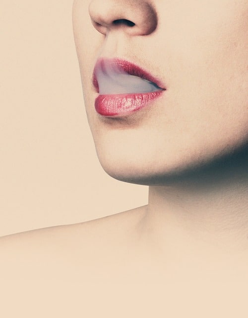Close-up of a person's lower face with pink lips slightly parted, exhaling a small cloud of vapour from e-cigs against a pale background.