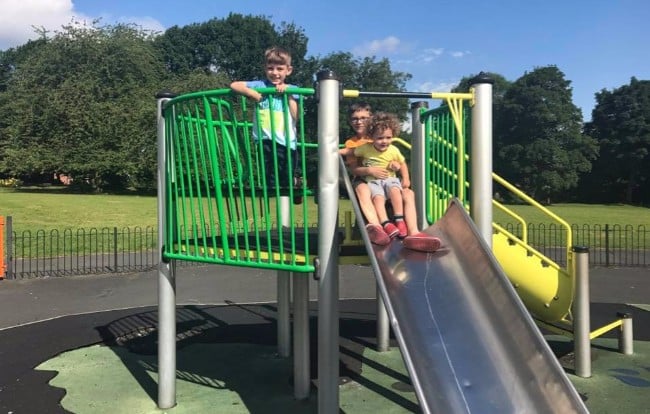 Three children play on a playground slide—a perfect example of affordable children’s entertainment. Two sit ready to go down, whilst one stands by the green railing. The outdoor playground is surrounded by trees and grass.