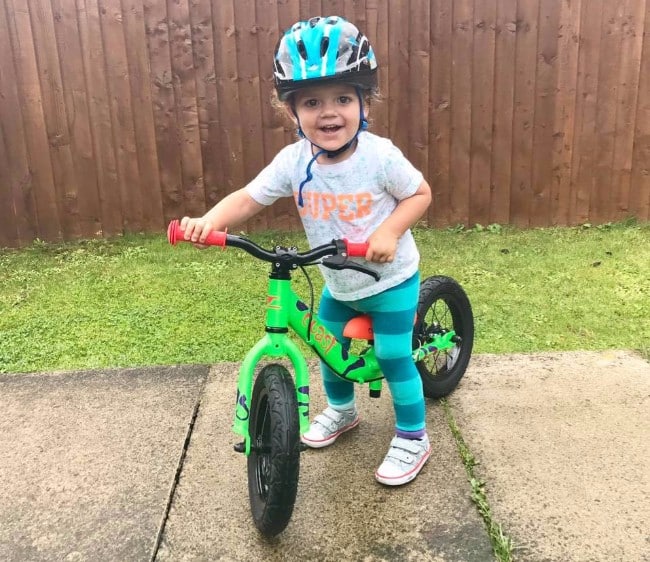 A smiling young child wearing a helmet, t-shirt, leggings, and trainers stands on a green balance bike—perfect for keeping children occupied with budget children's activities on the patio, with grass and a wooden fence in the background.