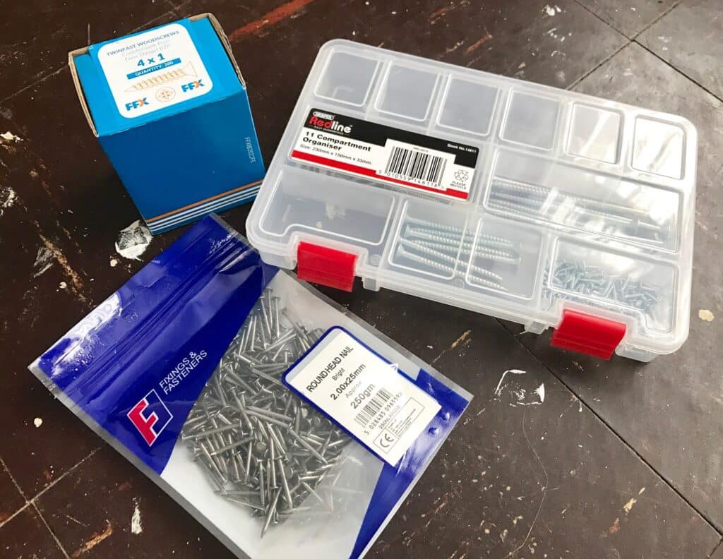 A packet of nails, a blue box of screws, and a clear plastic organiser with assorted screws are placed on a dark wooden floor—perfect for your DIY needs or when buying tools for your next project.
