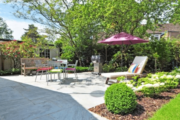 A sunny back garden patio with a wooden bench, metal table and chairs, colourful lounge cushions, a pink parasol, barbecue, and lush plants—perfect for relaxing or getting inspired by DIY garden projects and creative garden ideas.