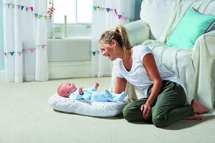 A woman smiles and leans towards a baby lying on a cushioned mat on the floor during the Aldi Baby & Toddler Event. They are in a bright, cosy living room with white curtains, bunting, and a sofa with colourful cushions.
