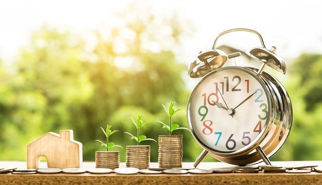 A wooden house model, three stacks of coins with growing plants, and a classic alarm clock are arranged on a table outdoors, illustrating estate planning and saving money for a child's inheritance over time.