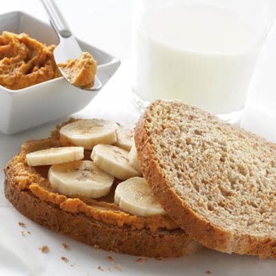A slice of wholegrain bread topped with nut butter and banana slices, perfect for easy lunch recipes, sits next to a plain slice of bread, a bowl of nut butter with a spoon, and a glass of milk in the background.