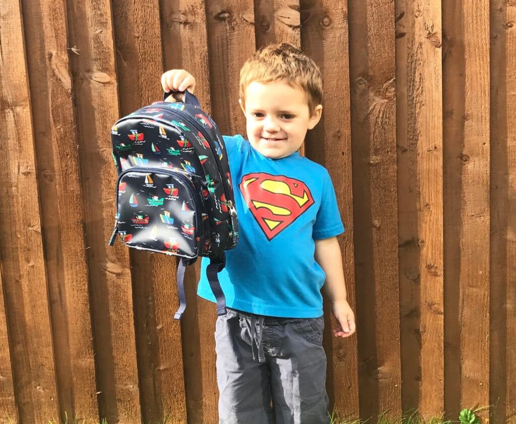 A young boy stands in front of a wooden fence, smiling and holding up a colourful rucksack filled with school supplies. He is wearing a blue T-shirt with a red and yellow Superman logo and grey trousers, ready for going back to school.