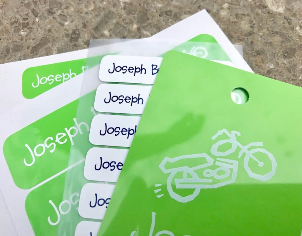 Green and white name labels reading "Joseph B" are spread out on a speckled worktop—perfect school supplies for going back to school. One green label features a white outline drawing of a motorbike, adding a fun touch to your back to school essentials.