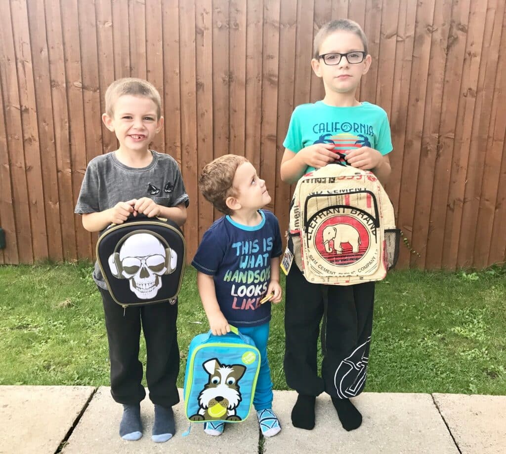 Three young boys stand in front of a wooden fence, each holding school supplies. The boy on the left has a skull lunchbox, the middle boy looks up at the tallest boy with glasses and an elephant-themed lunchbox—ready for back to school.