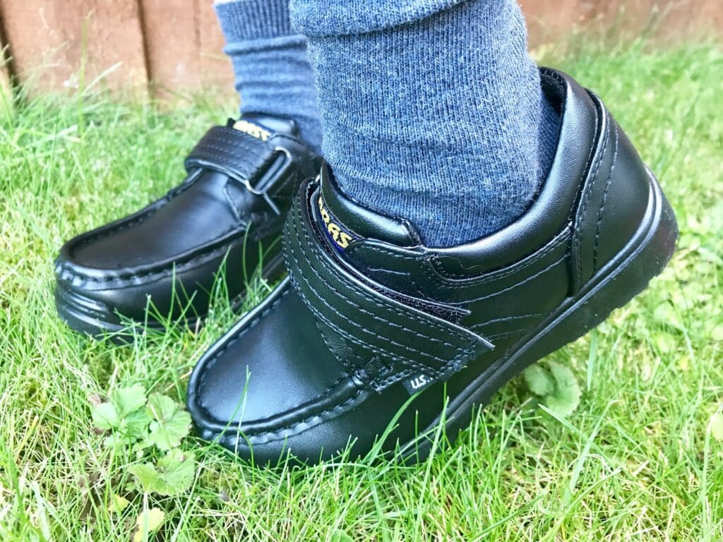 A close-up of a child wearing blue socks and black leather shoes with Velcro straps, standing on green grass outdoors—perfect for showcasing back to school essentials.