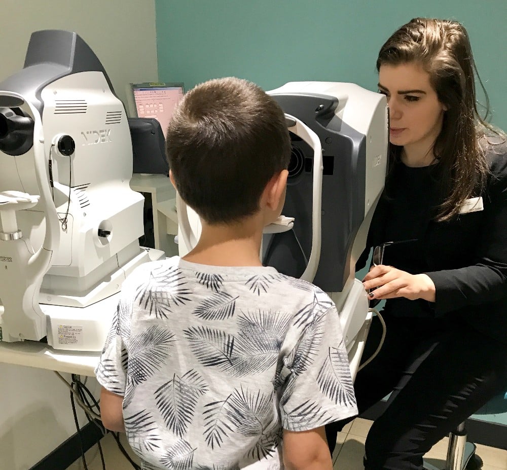 A young boy has his eyes examined with a Children’s Eye Test by a woman at Vision Express, who guides him as he sits facing the eye-testing machine in the clinic.