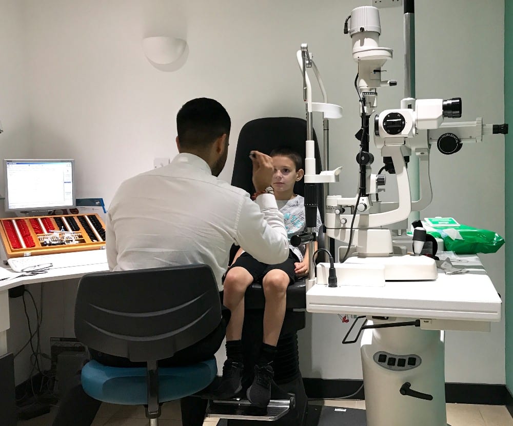 An optometrist performs a children's eye test on a young boy sitting in a chair, using specialised equipment in a bright, modern Vision Express examination room.