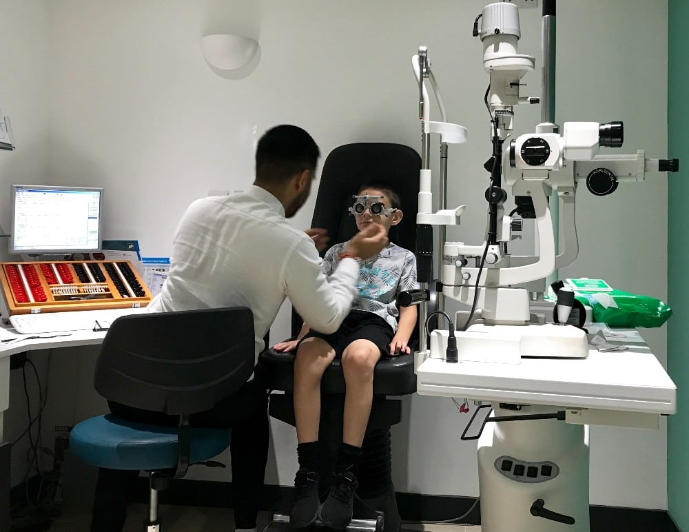 An optometrist at Vision Express conducts a Children's Eye Test, examining a young boy’s eyes with a phoropter in a modern clinic. The boy sits on a chair as the specialist adjusts the device, surrounded by medical equipment and a computer.