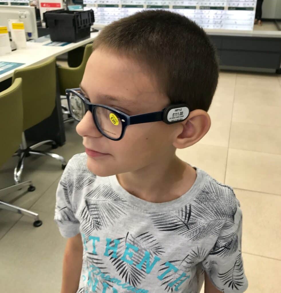 A young boy wearing blue glasses with a price tag still attached stands in what appears to be a Vision Express shop, perhaps after a children's eye test. He looks to the side and smiles slightly.
