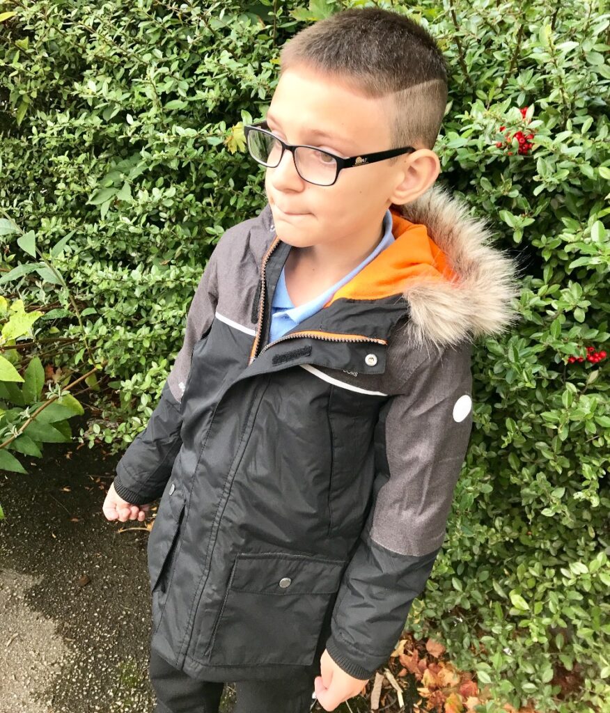 A young boy with short hair and glasses stands outside in front of green bushes, wearing a black and grey winter coat with an orange hood and fur trim. Ready for going back to school, he looks slightly to the side with a neutral expression.