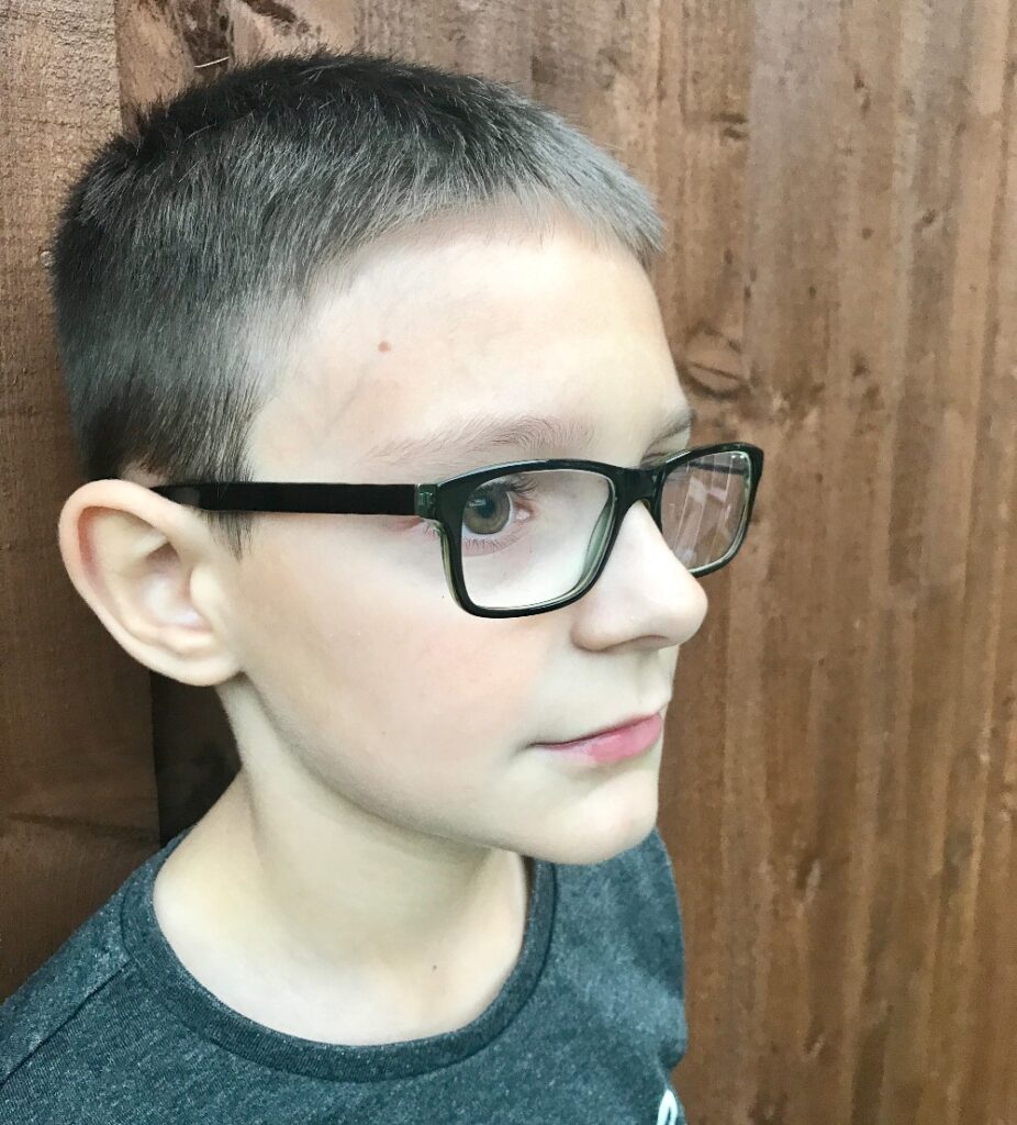 A young boy with short brown hair and black-framed glasses, fresh from a Children’s Eye Test at Vision Express, stands in front of a wooden fence looking to the right. He is wearing a dark grey shirt.