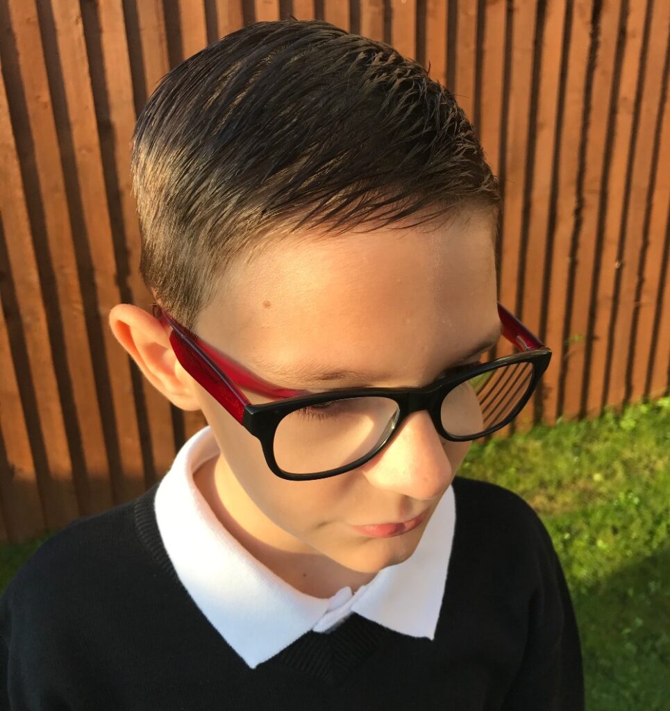 A young boy with neatly combed hair, wearing large black and red glasses from Vision Express, a white collared shirt, and a black jumper, stands outside near a wooden fence in sunlight after his children's eye test.