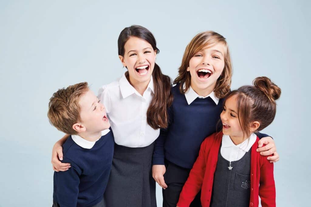 Four young children in school uniforms stand together, smiling and laughing. Two girls and two boys have their arms round each other against a light blue background, showing happiness and friendship as they get ready for going back to school.