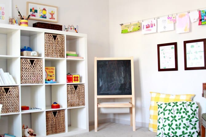 A bright, organised playroom doubles as a bedroom, featuring a white cubby shelf with baskets and toys, a black chalkboard easel, colourful cushions, framed children’s art on the wall, and more artwork hanging on a string.