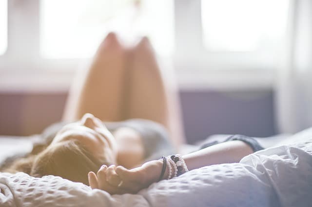A person is lying on their back on a bed, legs extended and slightly raised—a moment of self-care as sunlight streams through the window behind them, offering a peaceful pause for even the busiest parent. Their face is out of focus.