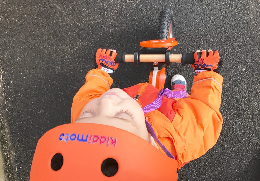 A young child wearing an orange helmet and jacket rides a Kiddimoto Scrambler Balance Bike on a paved surface, seen from above. The child’s hands grip the handlebars, and their gloves are orange with blue writing—ready for J's Adventures.