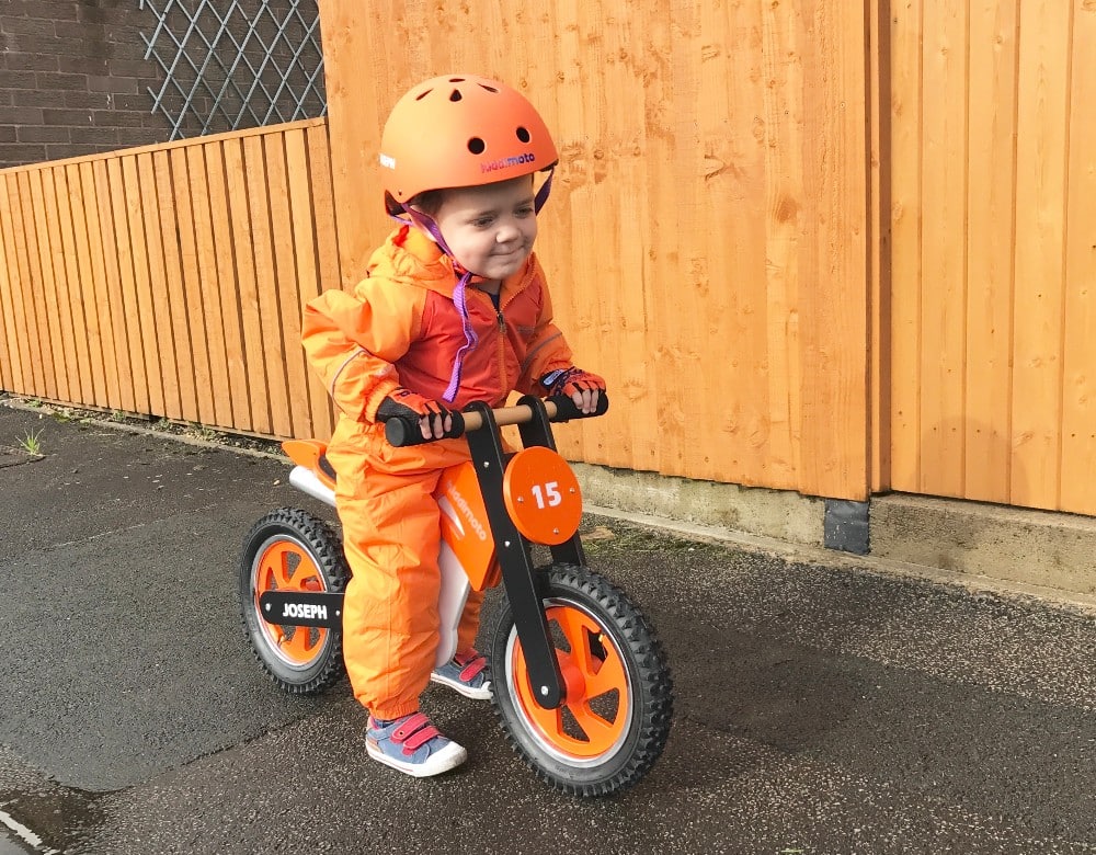 A young child wearing an orange helmet and jumpsuit rides a Kiddimoto Scrambler Balance Bike on wet pavement next to a wooden fence. Smiling, the child enjoys J's Adventures, with "Joseph" and the number 15 displayed on the bike.