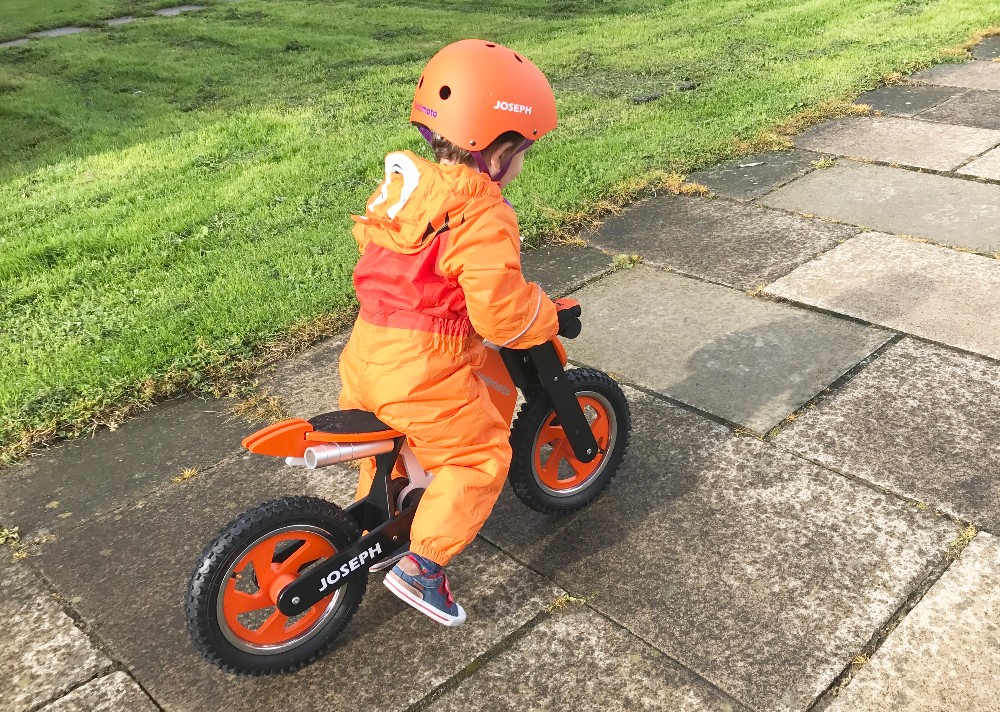 A young child in an orange helmet and outfit rides a Kiddimoto Scrambler Balance Bike on a stone pavement next to a grassy area.