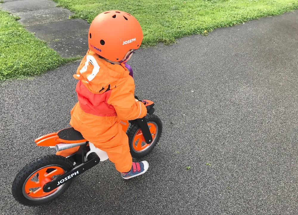 A young child wearing an orange helmet and jacket enjoys J's Adventures, riding a Kiddimoto Scrambler balance bike labelled "JOSEPH" on a paved path next to green grass.