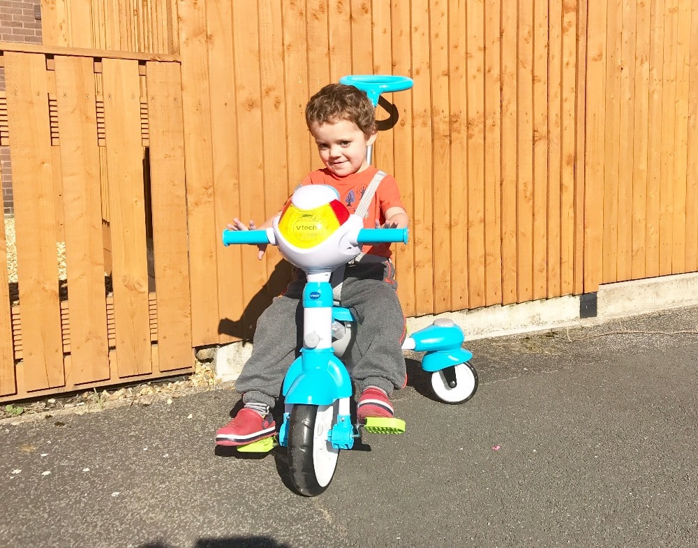 A young child wearing a red shirt and grey trousers rides a blue and white VTech tricycle on a paved surface, with a wooden fence and building in the background. The child looks happy and is smiling, enjoying their own Tour De Trike in Leeds.